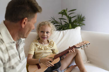 Smiling girl playing guitar sitting next to father in the living room at home - SVKF01672