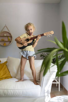 Glückliches Mädchen spielt Gitarre auf der Couch im Wohnzimmer zu Hause - SVKF01673