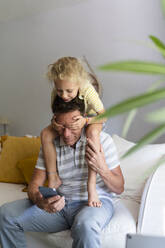 Girl sitting on father's shoulders and covering his eyes at home - SVKF01679
