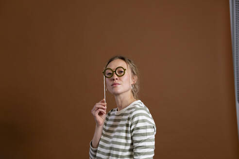 Confident woman holding eyeglasses prop over brown background - MIKF00397