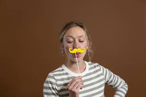 Woman puckering and holding mustache prop against brown background - MIKF00400