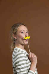 Blonde Frau hält Schnurrbartstütze vor braunem Hintergrund - MIKF00403