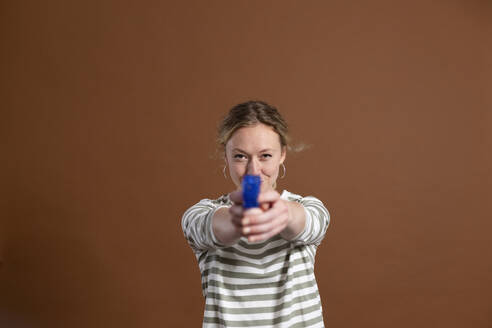 Blonde Frau hält Spielzeugpistole auf braunem Hintergrund - MIKF00409