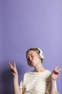 Frau mit Kopfhörern gestikuliert Friedenszeichen gegen lila Hintergrund - MIKF00424