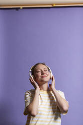 Lächelnde Frau mit geschlossenen Augen hört Musik auf Kopfhörern vor lila Hintergrund - MIKF00428