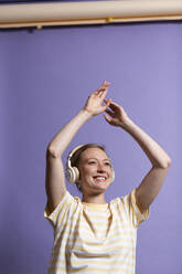 Lächelnde Frau hört Musik über Kopfhörer und tanzt auf lila Hintergrund - MIKF00429