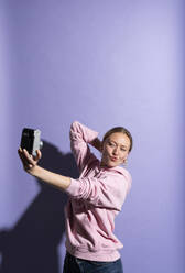 Frau schmiegt sich an und fotografiert sich selbst durch die Kamera vor lila Hintergrund - MIKF00434