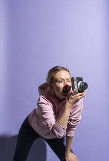 Frau beugt sich und fotografiert durch die Kamera vor lila Hintergrund - MIKF00436