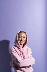 Smiling woman standing with arms crossed against purple background - MIKF00440