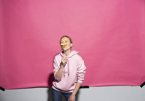 Verspielte Frau hält Schnurrbart-Requisite vor rosa Hintergrund - MIKF00446