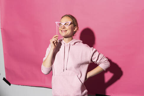 Lächelnde Frau mit Hand auf der Hüfte, die eine Brille vor einem rosa Hintergrund hält - MIKF00449