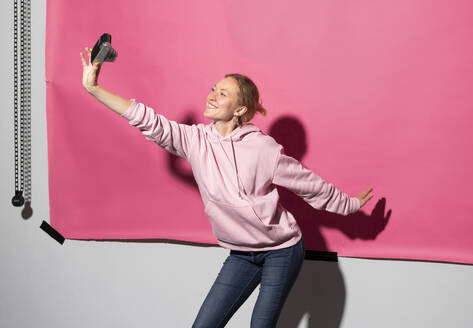Happy woman taking selfie through camera against pink background - MIKF00452