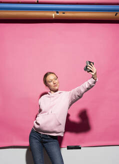 Woman in hooded shirt taking selfie with camera over pink background - MIKF00453