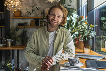 Lächelnder junger Geschäftsmann, der sich in einem Café an einen Tisch lehnt - VPIF08736