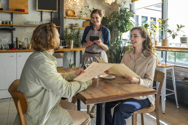 Kellnerin im Gespräch mit zufriedenen Kunden, die mit Menükarten in einem Café sitzen - VPIF08745