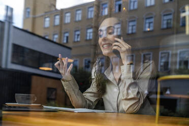 Smiling businesswoman talking on mobile phone at cafe seen through glass - VPIF08767