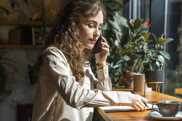Junge Geschäftsfrau, die mit einem Smartphone telefoniert und Dokumente auf dem Tisch eines Cafés liegen hat - VPIF08769