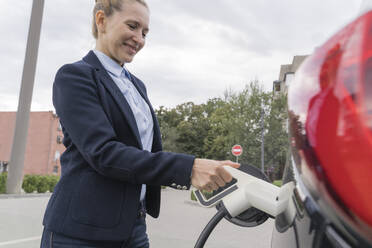 Lächelnde Geschäftsfrau beim Aufladen eines Elektroautos an einer Tankstelle - OSF02194