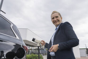 Smiling businesswoman holding charger next to electric car at station - OSF02195