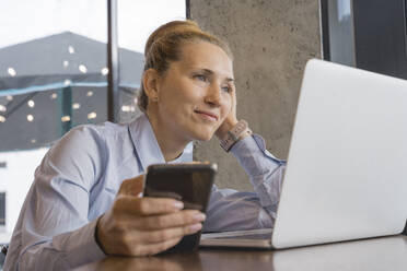 Lächelnde Geschäftsfrau mit Smartphone, die sich in einem Café auf einen Tisch mit Laptop stützt - OSF02198