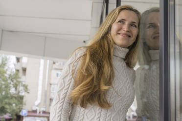 Happy woman wearing sweater and leaning on glass door - OSF02202