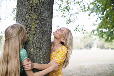 Mother and daughter hugging tree trunk in park - AMWF01865