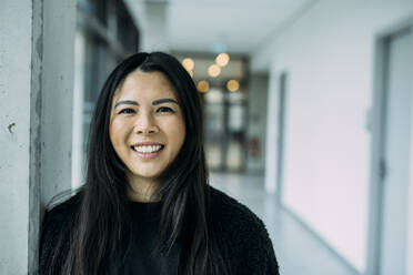 Smiling beautiful businesswoman with long black hair at office corridor - JOSEF21312