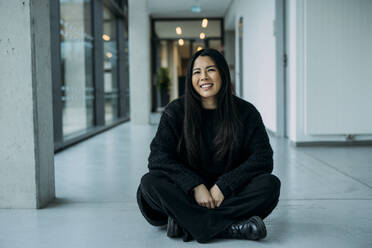 Smiling beautiful businesswoman sitting cross-legged on floor in corridor at workplace - JOSEF21325