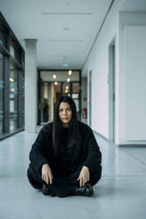 Confident businesswoman sitting cross-legged on floor in corridor at workplace - JOSEF21326