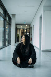 Lächelnde Geschäftsfrau, die im Schneidersitz auf dem Boden eines Korridors am Arbeitsplatz sitzt - JOSEF21328