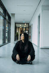 Smiling businesswoman sitting cross-legged on floor in corridor at workplace - JOSEF21328