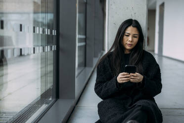 Geschäftsfrau, die sich an eine Säule lehnt und ihr Smartphone im Korridor am Arbeitsplatz benutzt - JOSEF21336