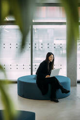 Happy businesswoman sitting on seat with laptop in office - JOSEF21351