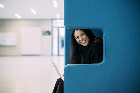 Lächelnde schöne Geschäftsfrau sitzt mit Laptop auf blauem Stuhl am Arbeitsplatz - JOSEF21353