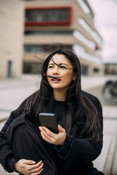 Geschäftsfrau mit Smartphone vor Bürogebäude sitzend - JOSEF21366