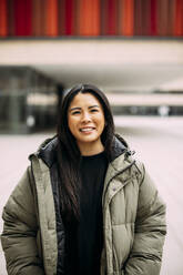 Smiling businesswoman wearing padded jacket outside office building - JOSEF21369