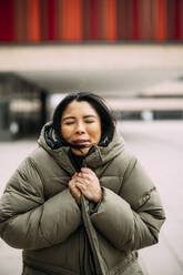 Geschäftsfrau mit geschlossenen Augen in gepolsterter Jacke vor einem Bürogebäude - JOSEF21370