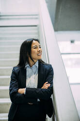 Smiling beautiful businesswoman with arms crossed leaning on staircase railing at office - JOSEF21377