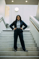 Happy beautiful businesswoman with arms akimbo standing on staircase at office - JOSEF21382