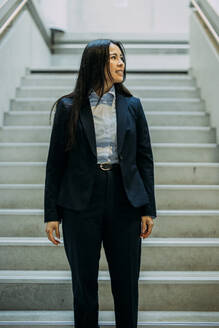 Lächelnde Geschäftsfrau, die auf einer Treppe im Büro steht und wegschaut - JOSEF21383