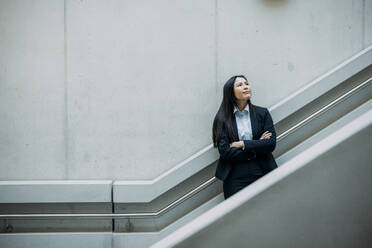 Businesswoman with arms crossed leaning on staircase railing at workplace - JOSEF21385