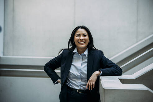 Happy businesswoman leaning on staircase railing at workplace - JOSEF21386