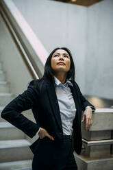 Beautiful businesswoman leaning on staircase railing looking up at office - JOSEF21387