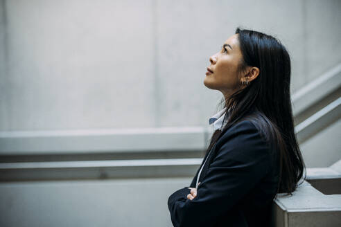 Businesswoman looking up with arms crossed leaning on railing at workplace - JOSEF21393