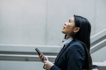 Businesswoman with smart phone looking up at workplace - JOSEF21397