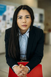 Selbstbewusste schöne Geschäftsfrau mit verschränkten Händen, die sich auf einen roten Stuhl im Büro stützt - JOSEF21406