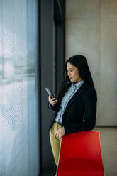 Businesswoman using smart phone leaning on red chair by window at office - JOSEF21410