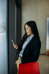 Businesswoman using smart phone leaning on chair at office - JOSEF21411
