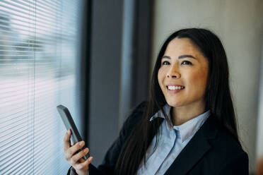 Smiling businesswoman using smart phone by window at office - JOSEF21413