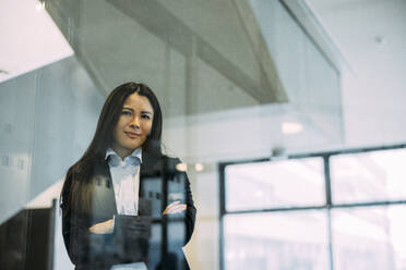 Confident businesswoman with arms crossed seen through glass at workplace - JOSEF21418